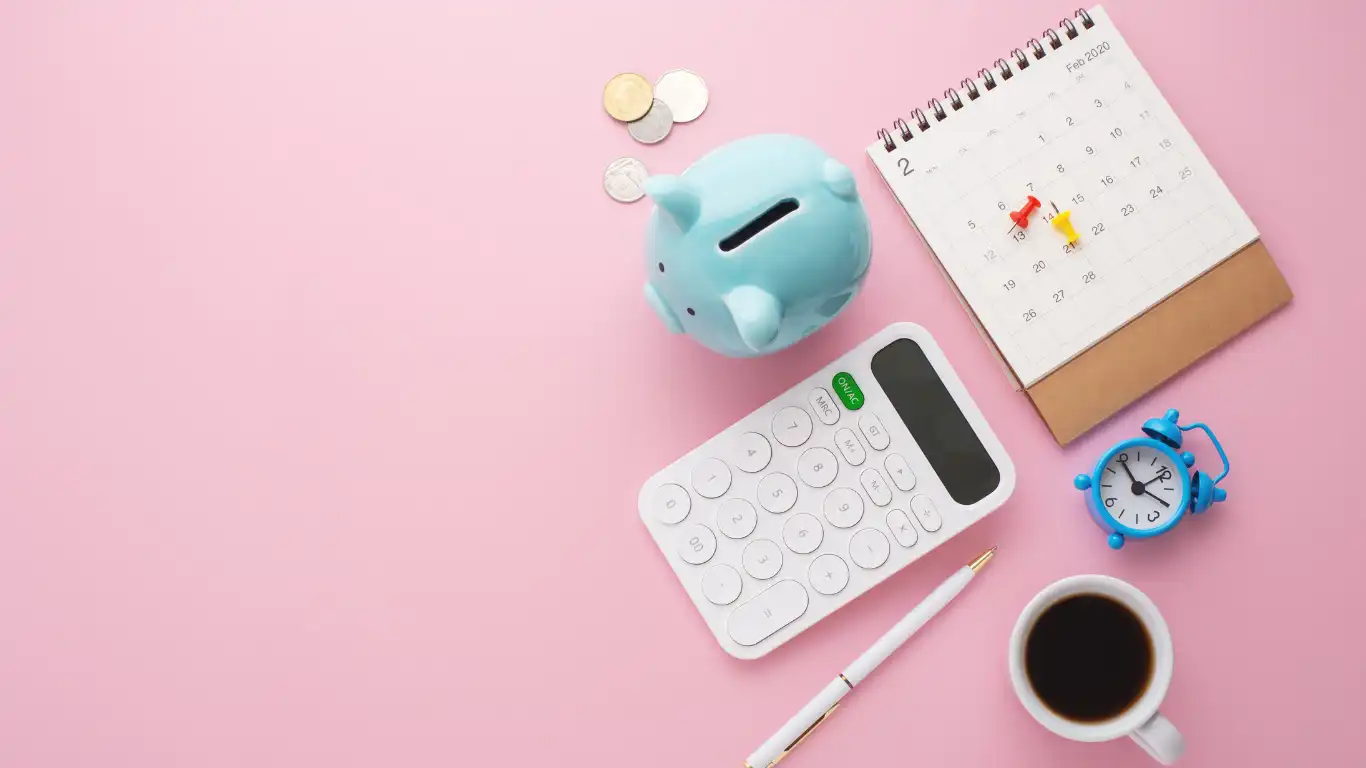ピンクの机の上に置かれた貯金箱、電卓、カレンダー、時計、ボールペン、コーヒーカップ